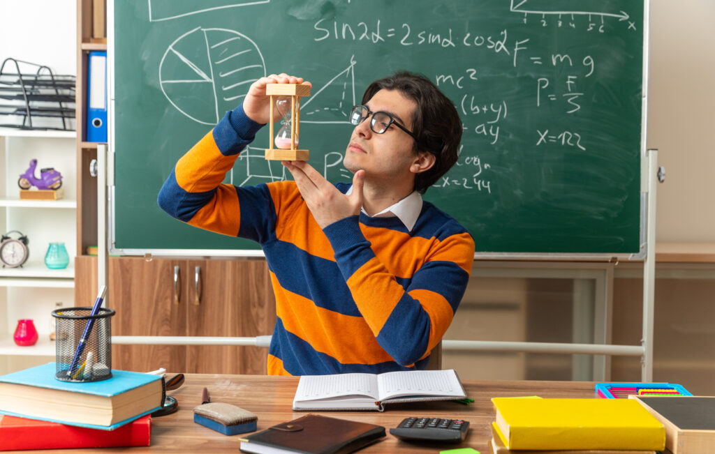 serious young caucasian geometry teacher wearing glasses sitting at desk with school tools in classroom holding hourglass vertically looking at it